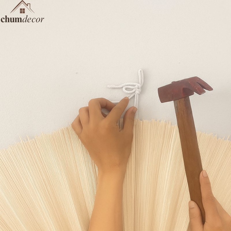 Hands preparing to hang an ivory bamboo fan wall using a white cord and hammer